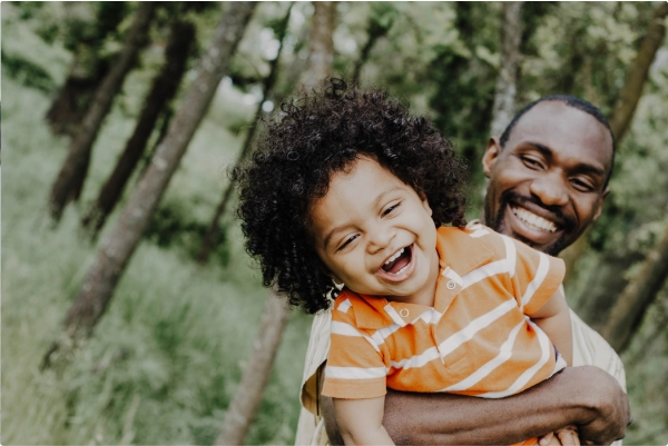 A man smiling and carrying a young child in a field with trees.