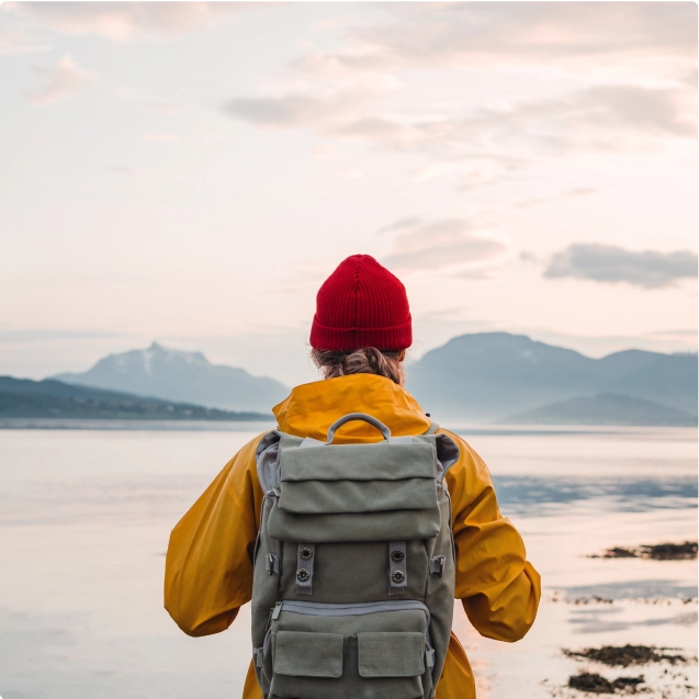 A person wearing a red hat and yellow jacket, looking out over a mountain lake.